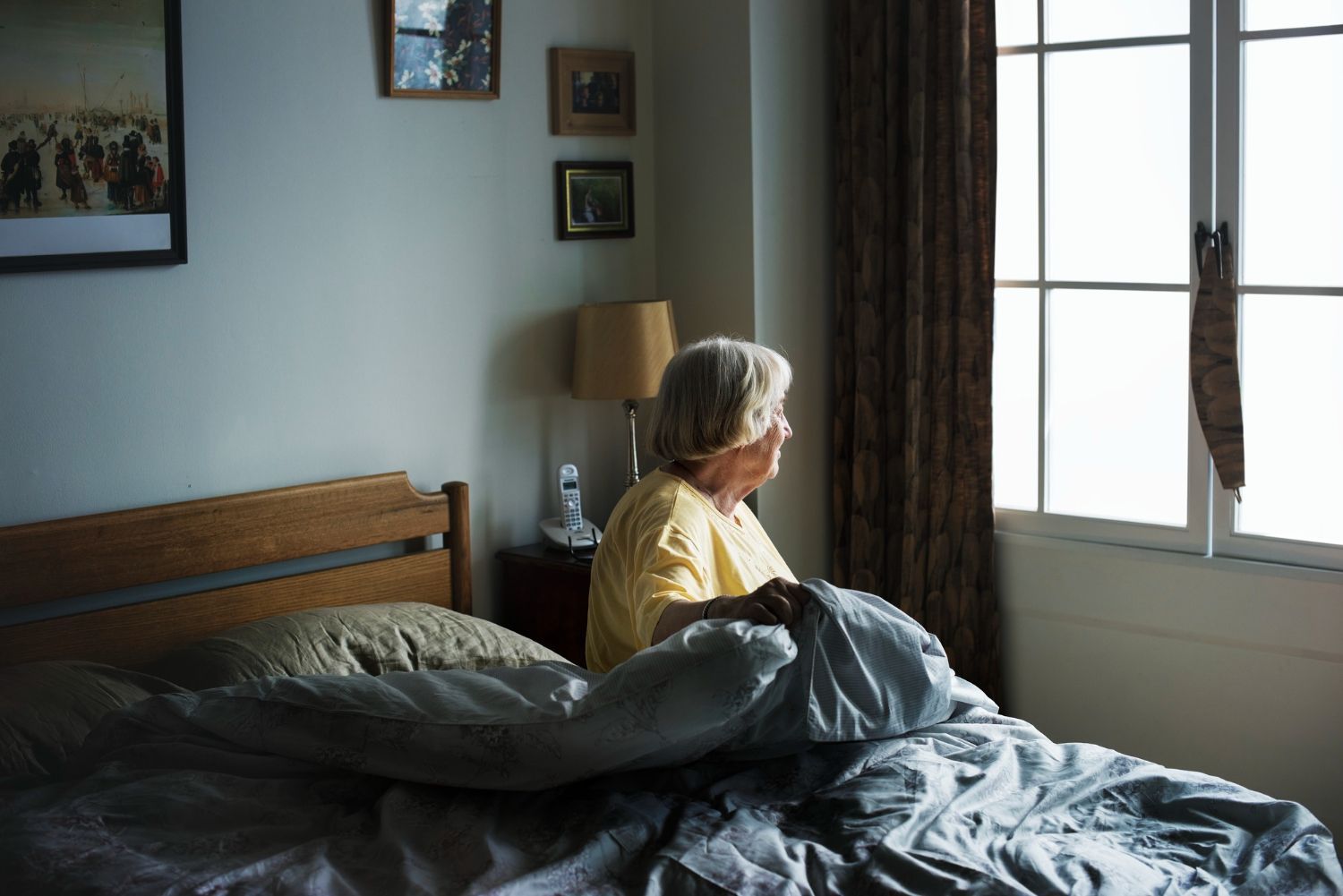 Woman sits on a bed, adjusting a blanket near a window with sunlight, bedside lamp, and framed art.