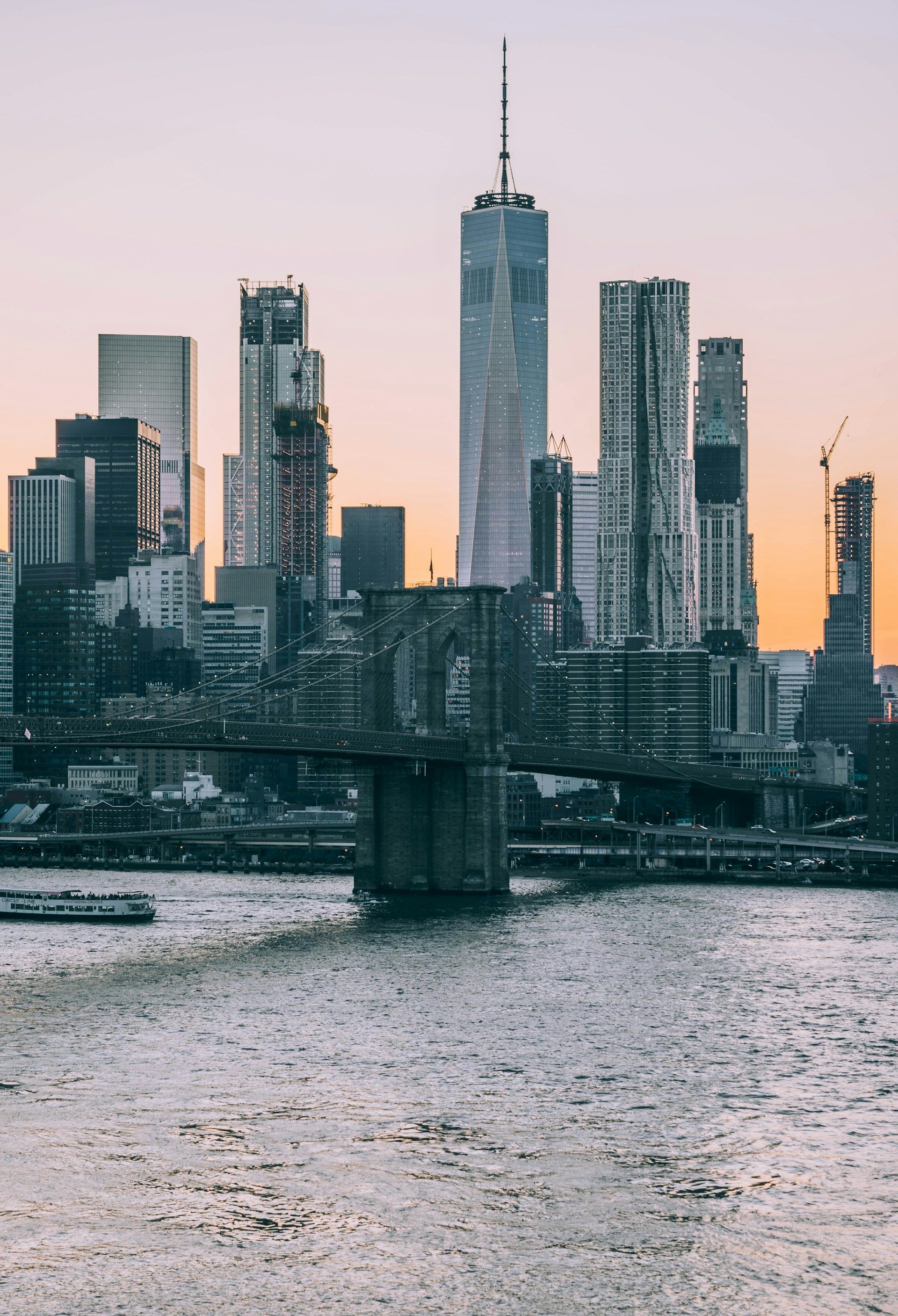 New York City skyline at dusk, including the Brooklyn Bridge and One World Trade Center over the water.