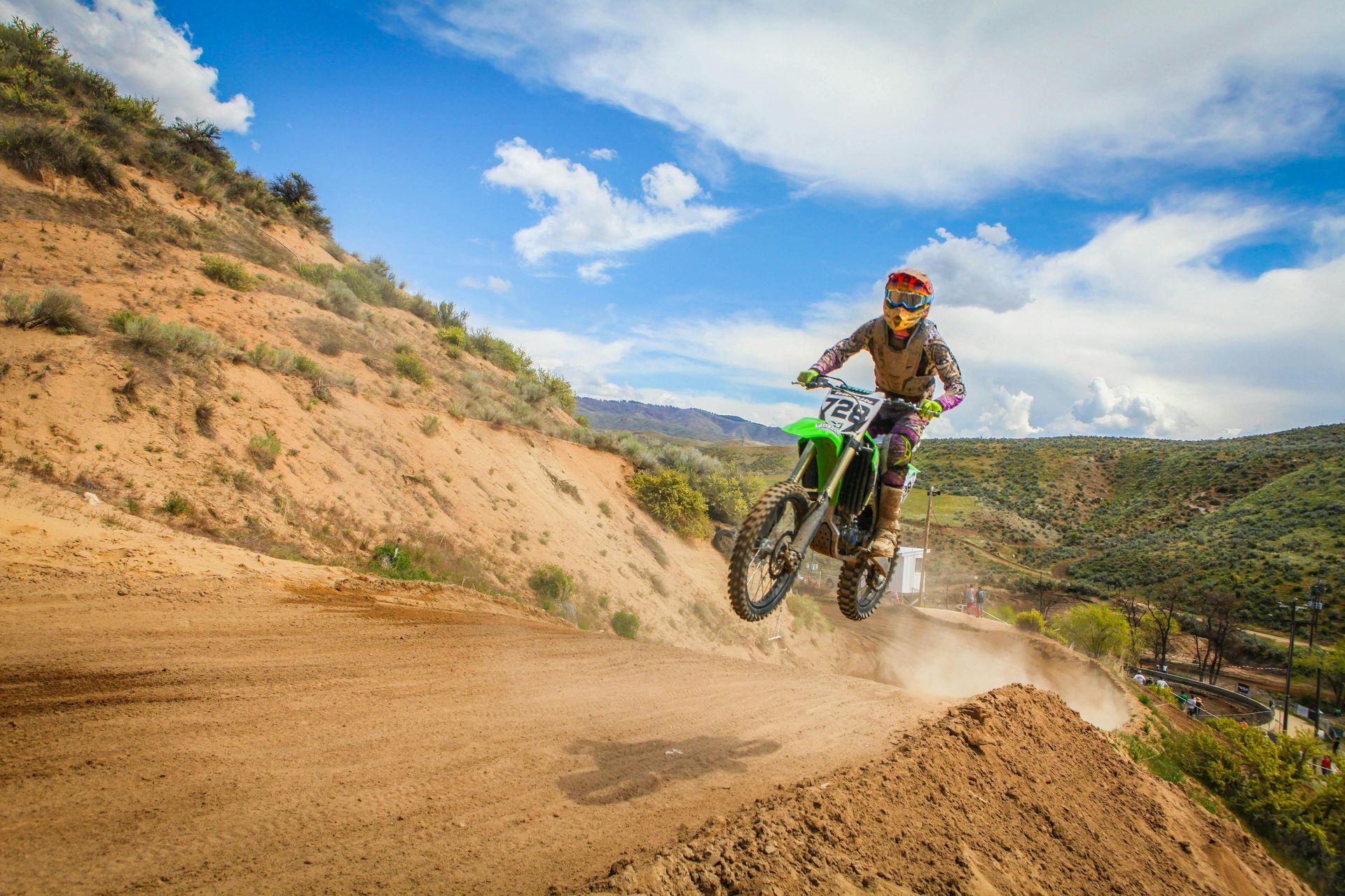 Dirt bike rider in mid-air, jumping over a dirt mound. Brown and green landscape with a blue sky.