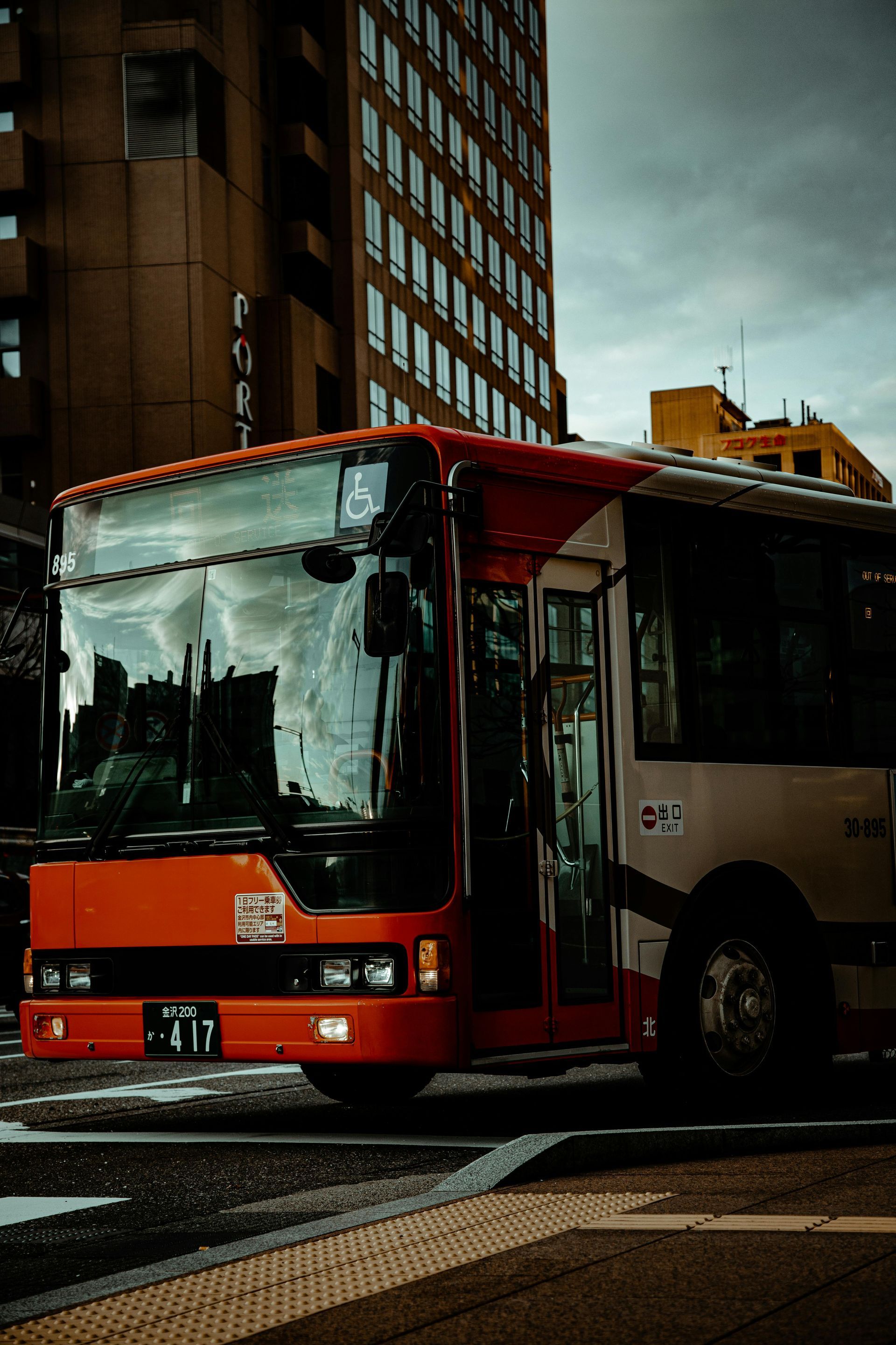 Red and white bus with open door on a city street, tall buildings and overcast sky.