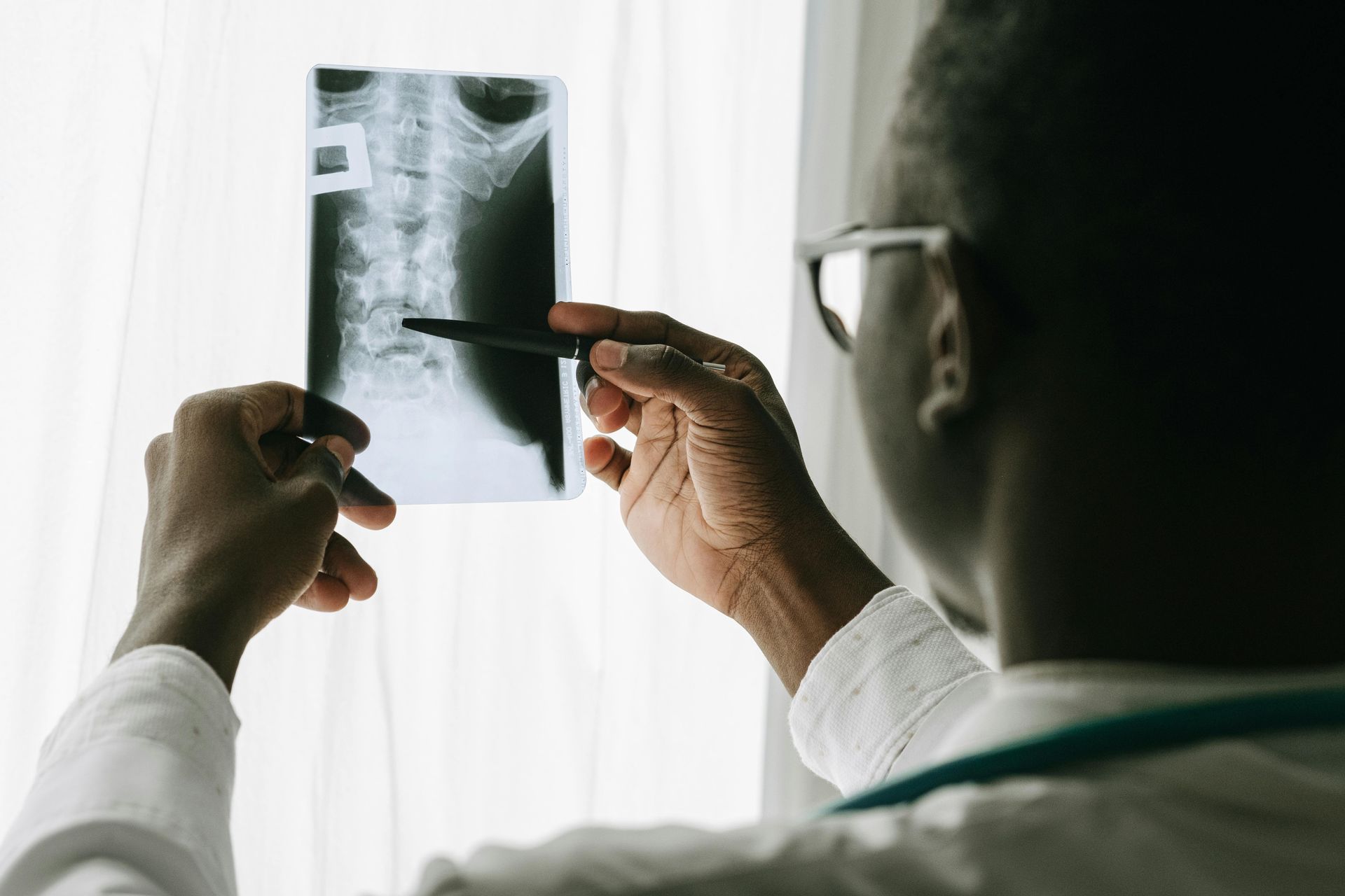 Doctor examines spinal X-ray with a pen, standing near a window.