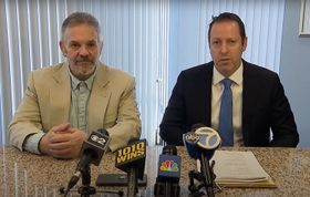 Two men seated at a table with microphones, likely for a press conference. One in a suit, the other in a blazer.