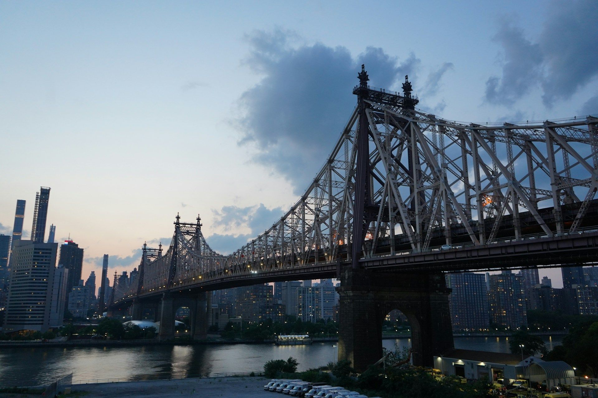 Queensboro Bridge at dusk over water, with city skyline in the background under a cloudy sky.