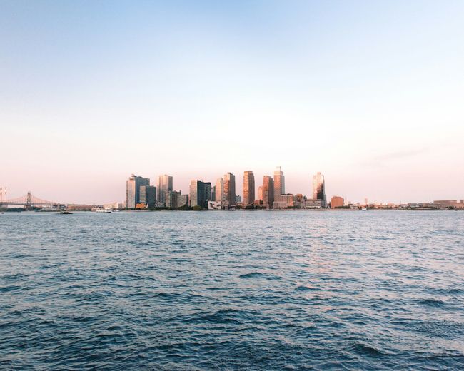 Skyline of city buildings across blue water under a clear, pale sky.