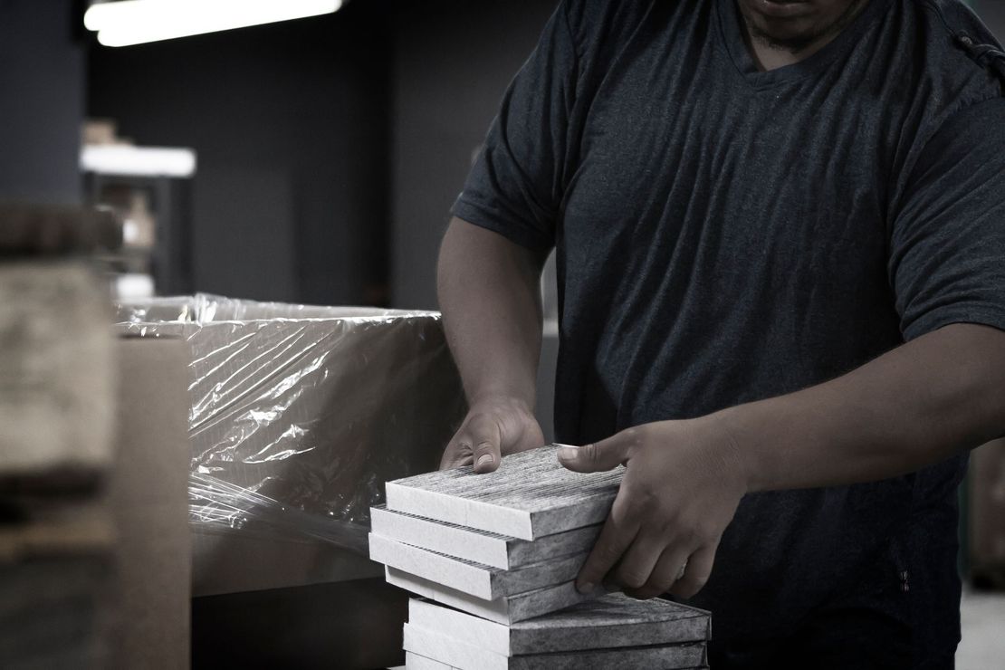 Person in a gray shirt stacks gray and white tiles by a wrapped cardboard box.