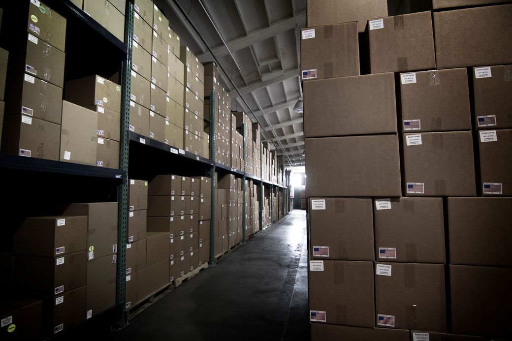 Warehouse interior with rows of stacked cardboard boxes on shelves and floor.