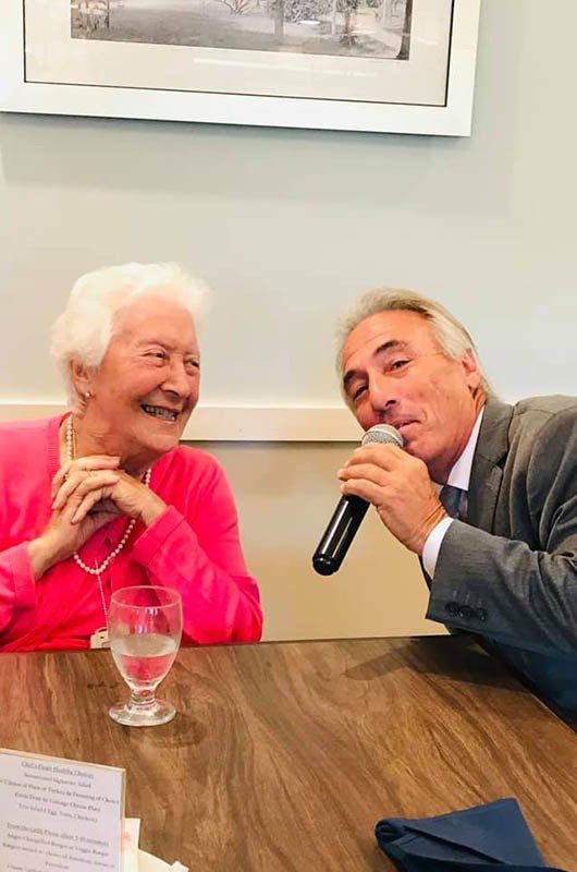 A man is singing into a microphone while sitting next to an older woman at a table.