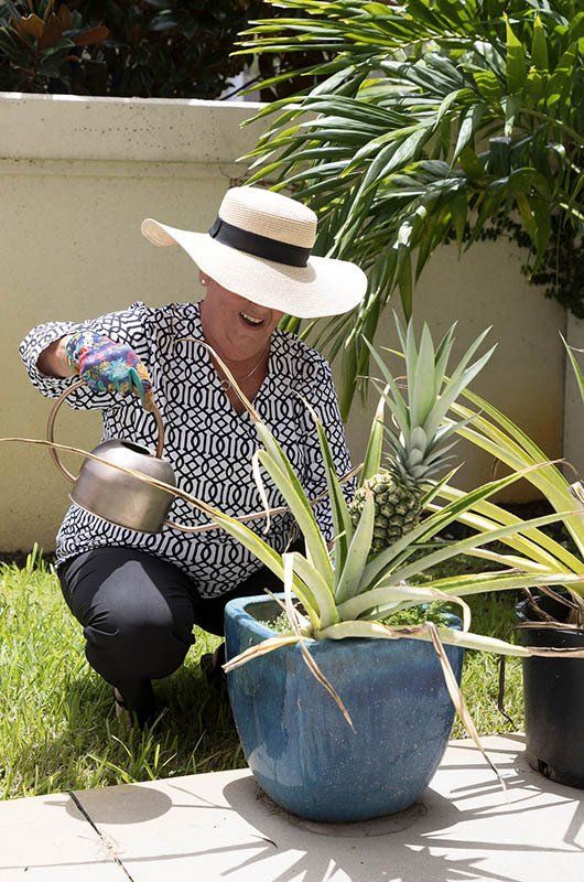 A woman in a hat is watering a pineapple in a blue pot.