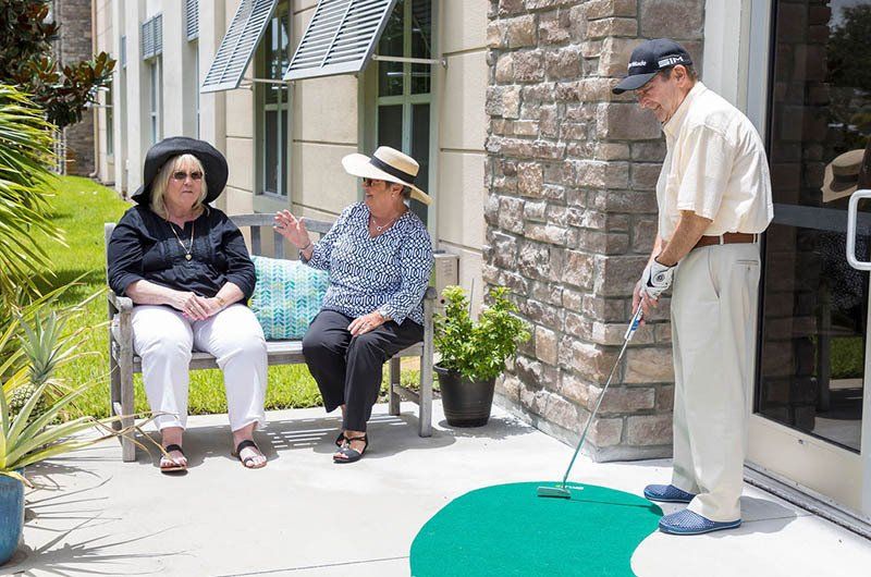 A man is putting a golf ball on a green mat while two women sit on a bench.