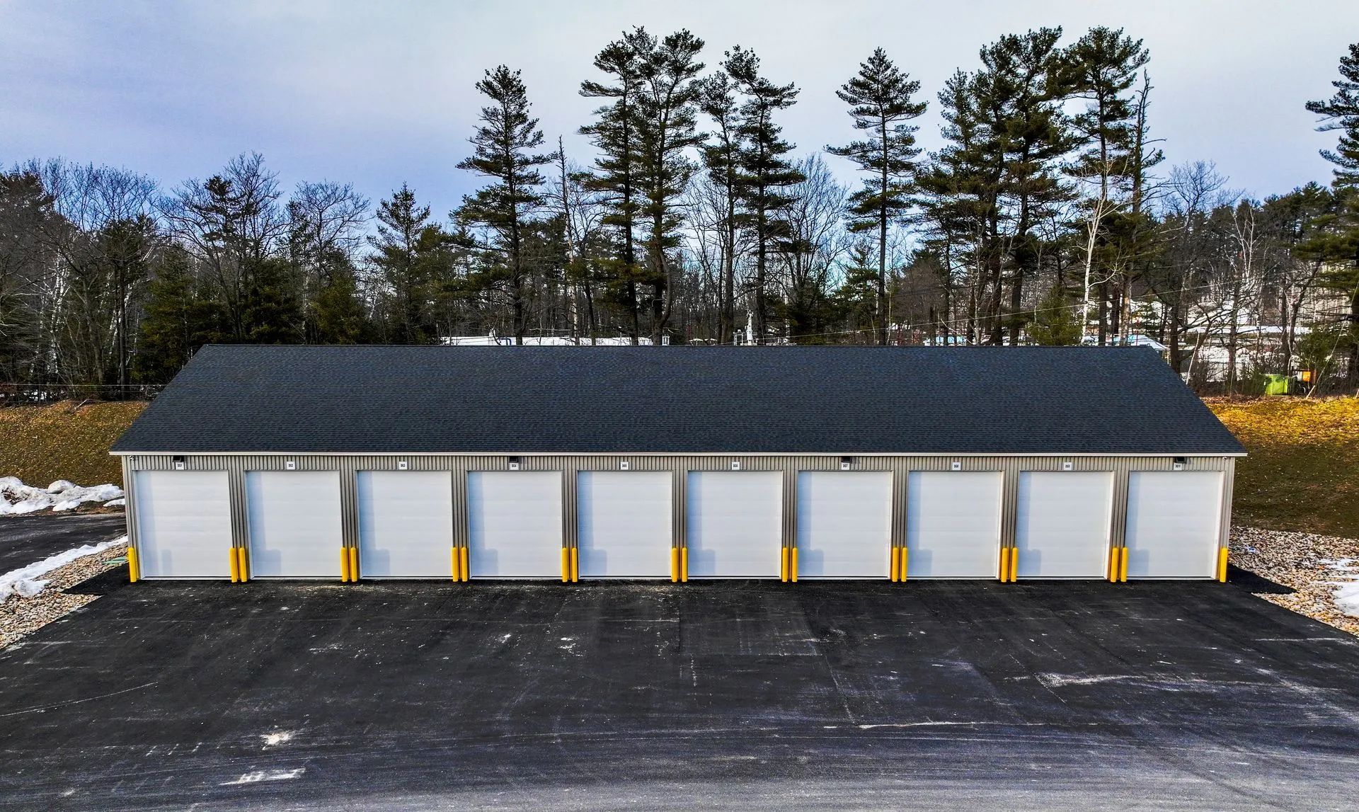 Storage units with gray doors and black roof, set against a backdrop of trees and a cloudy sky.