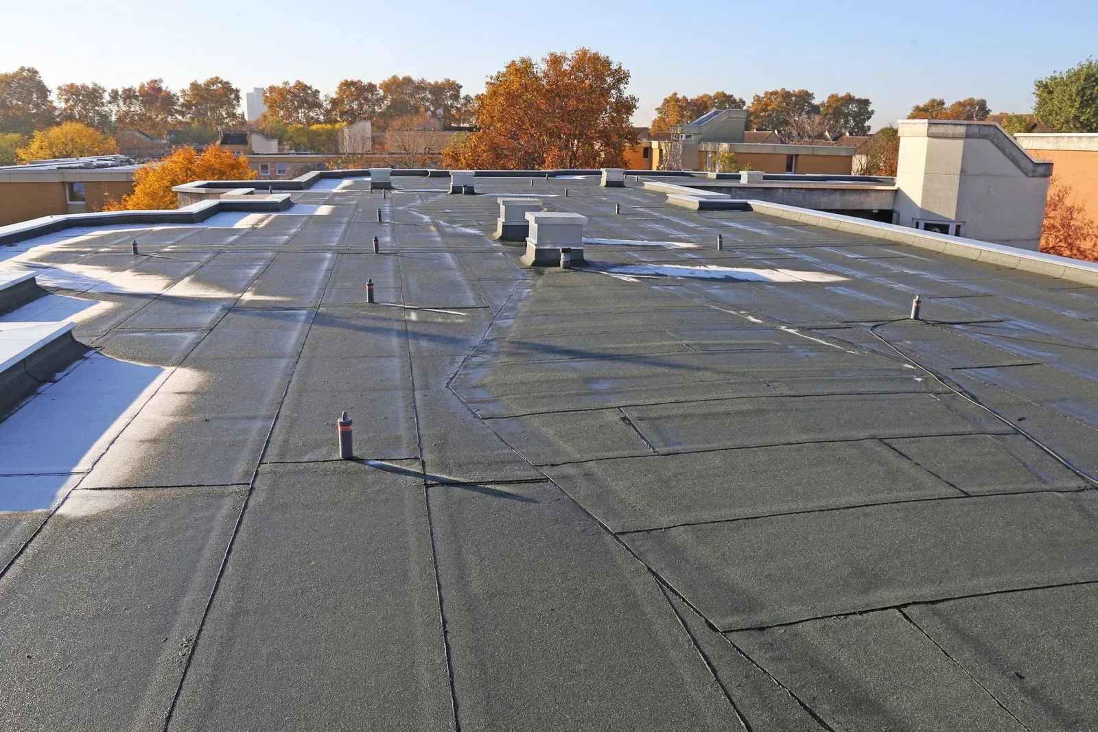 Flat commercial roof with dark material, patches of snow, and vents on a sunny day.