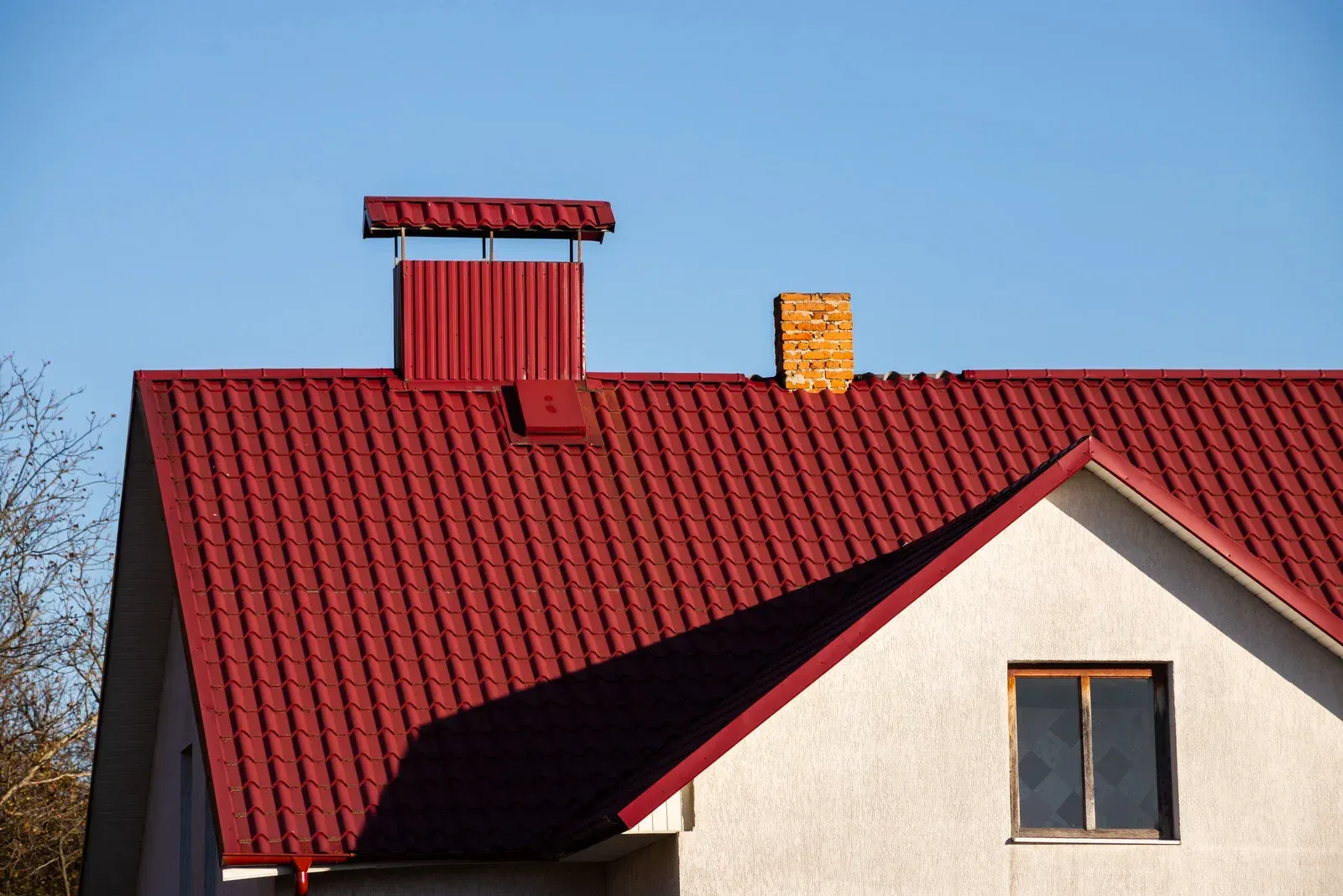 Red metal roof on a white house with a chimney against a blue sky.