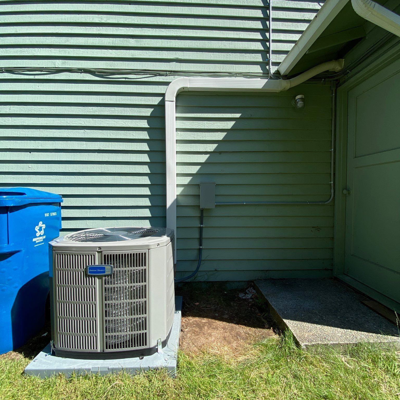 Air conditioning unit outside a green building with a blue trash bin.