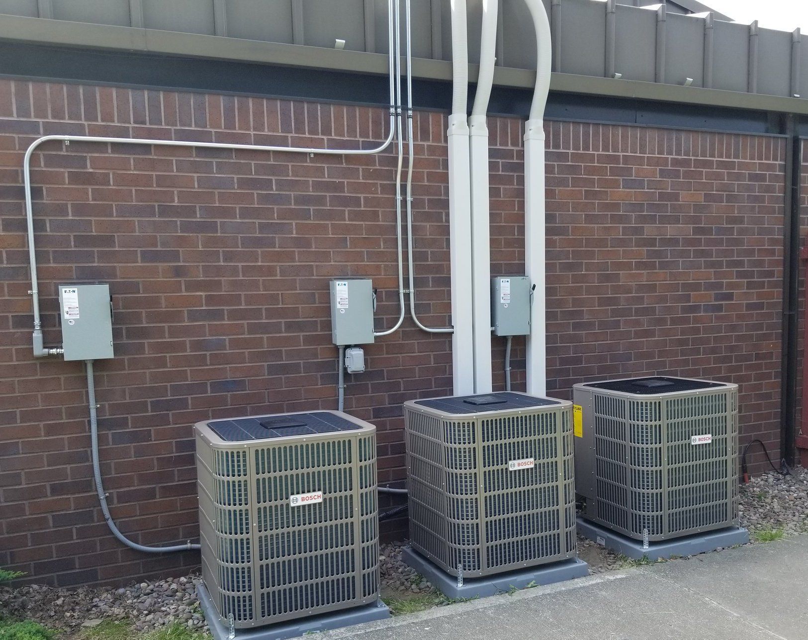 Three air conditioning units against a brick wall, with electrical conduits and junction boxes.