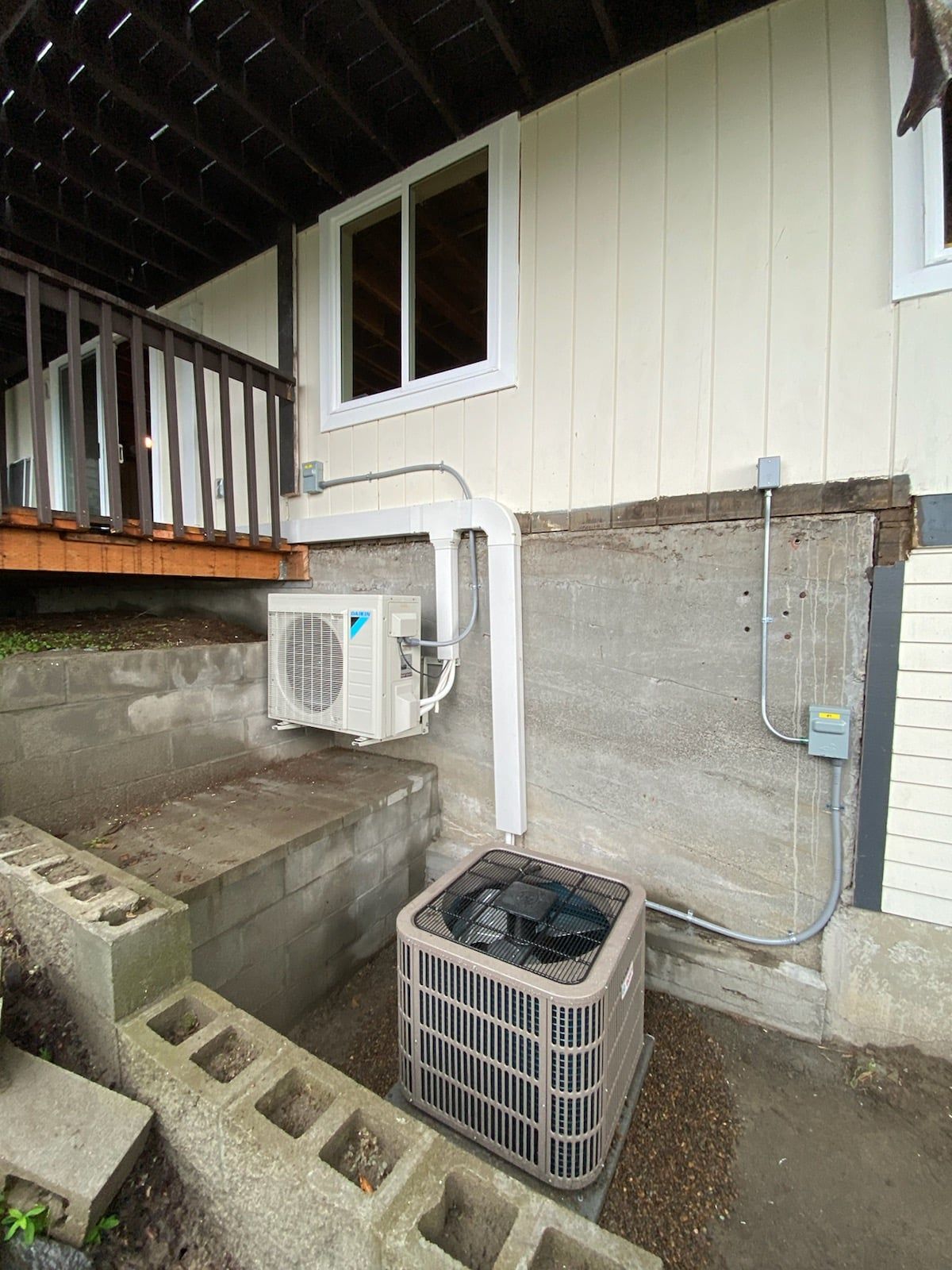 Outdoor HVAC units on a concrete wall. One is mounted, the other is ground-level, with connecting pipes.