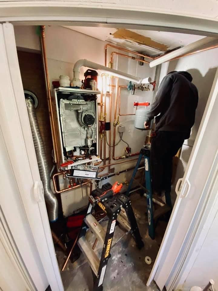 Person working on a heating system in a utility room. Copper pipes, tools, and a ladder are visible.