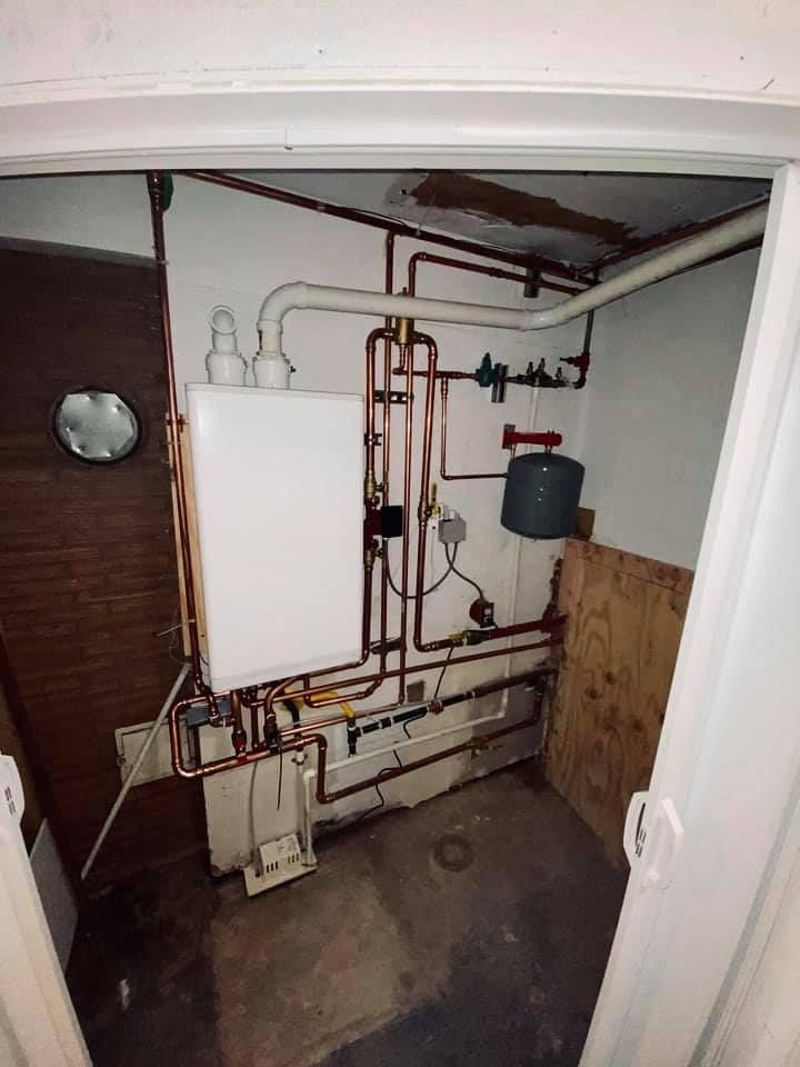 White-walled utility closet with a boiler, copper pipes, and a small water tank.