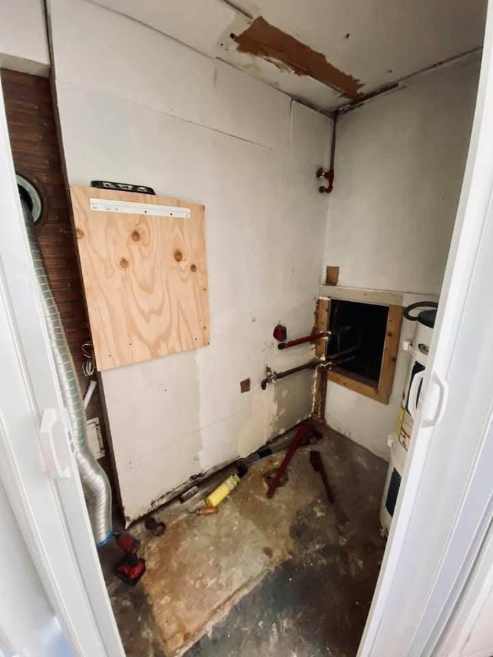 A small, unfinished utility room with exposed pipes, a wooden panel, and a partially open vent.
