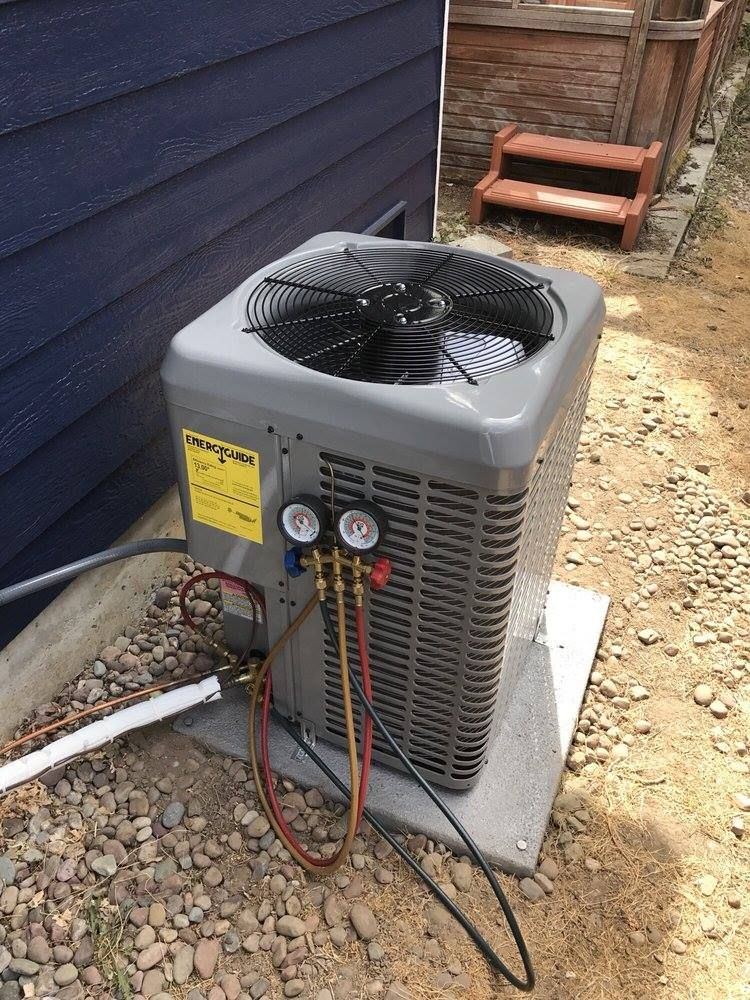 An air conditioning unit with gauges connected, set on a concrete pad next to a blue wall and gravel.