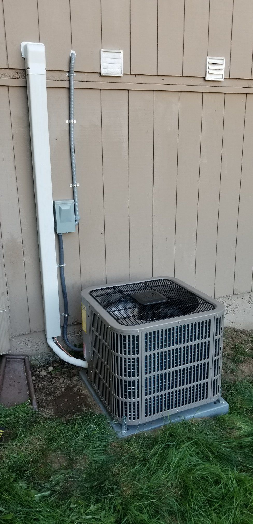 An air conditioning unit next to a light-colored fence with a white pipe running up it and vent-like structures.