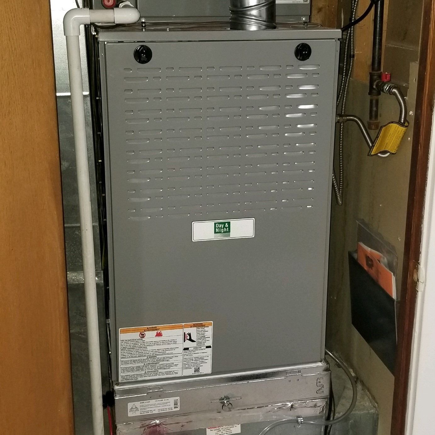 Gray furnace with labels, installed against a wall, white piping on left.