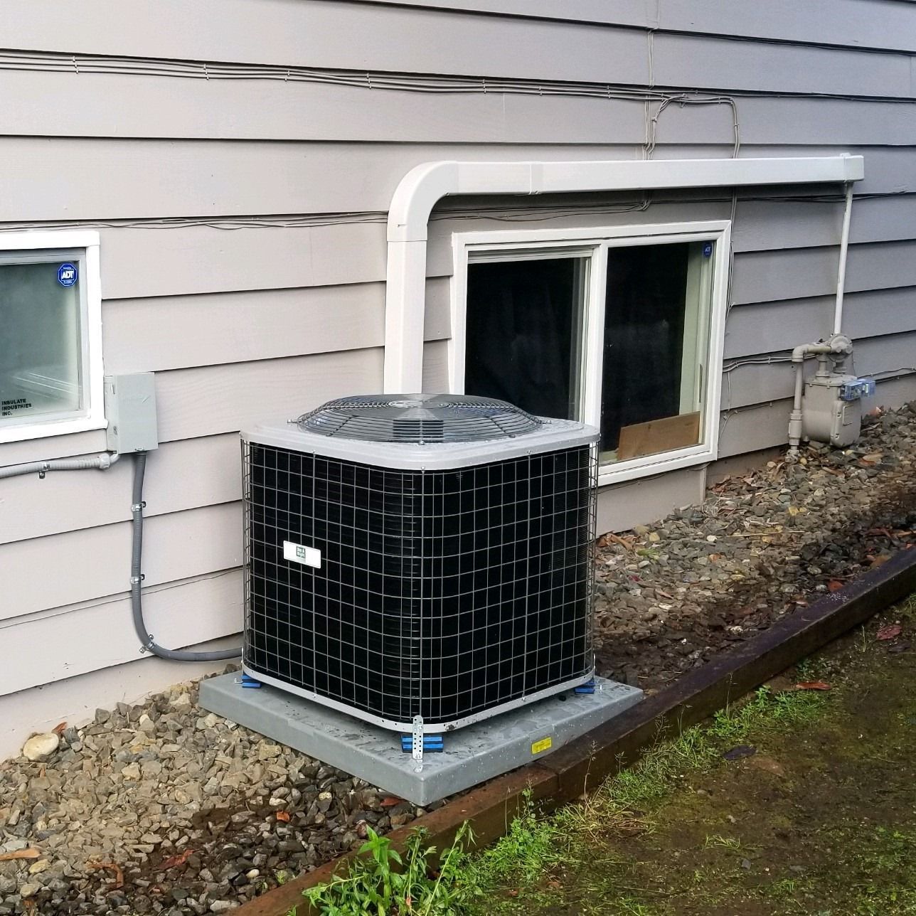AC unit on a gray base next to a house with a window, gas meter, and siding.