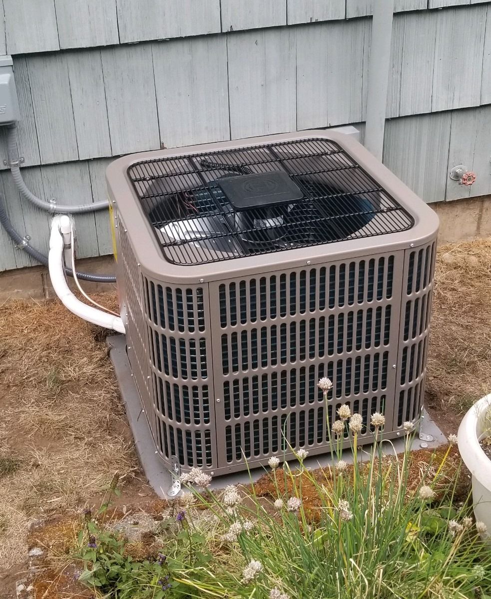 Air conditioning unit outside a light-colored house. Brown metal exterior with black top, sits on a concrete pad.