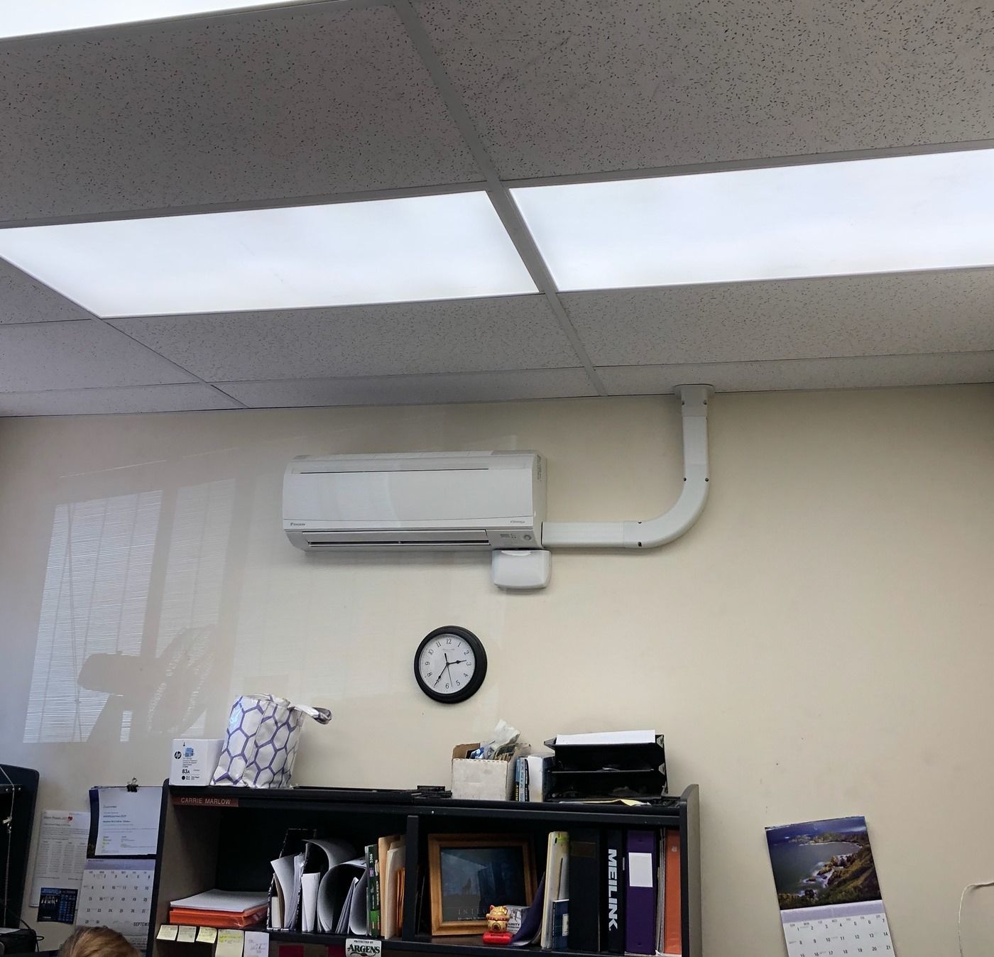 White wall-mounted air conditioner with piping, above a clock and office shelving.