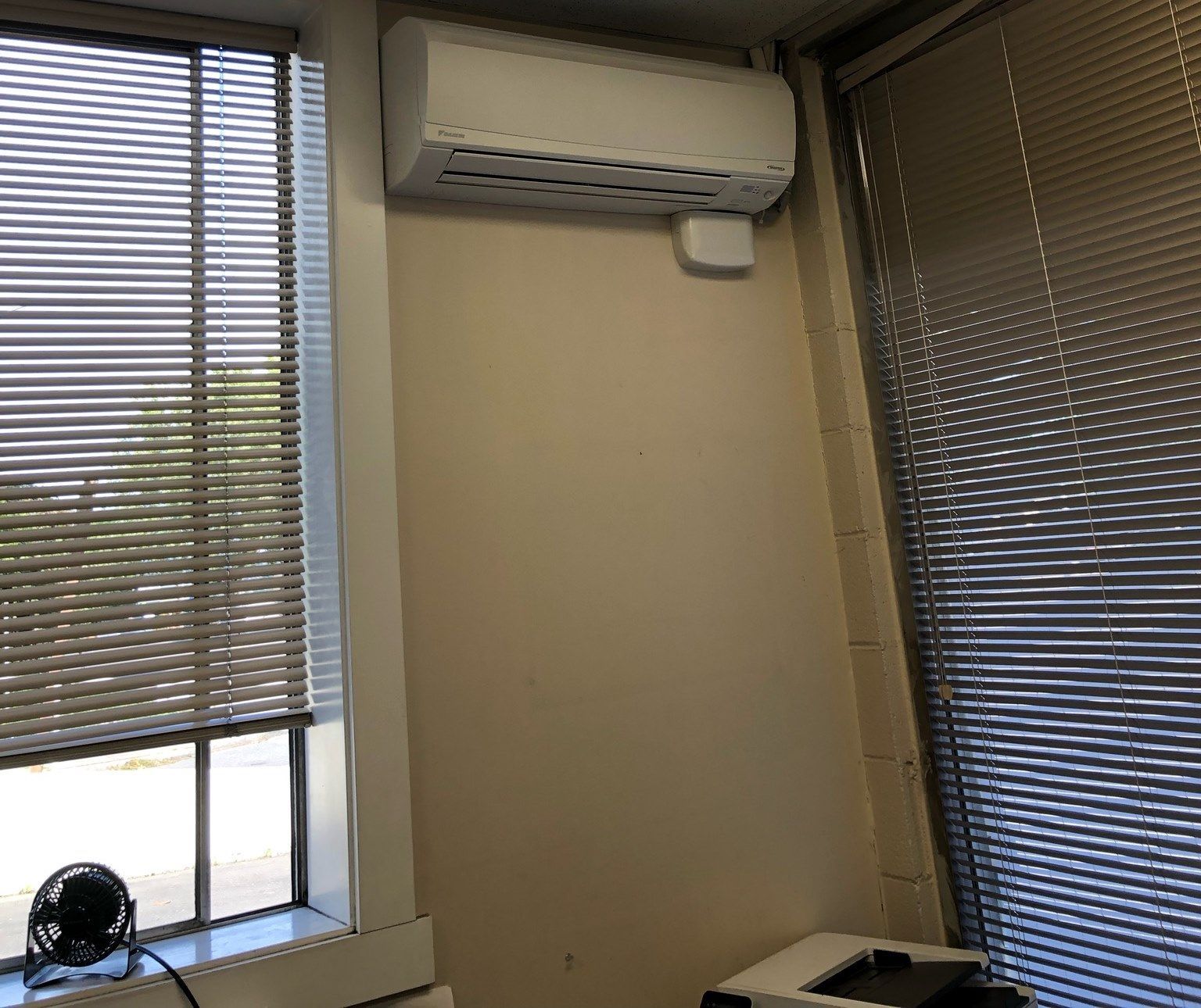 An indoor air conditioning unit is mounted on a beige wall next to a window with blinds.