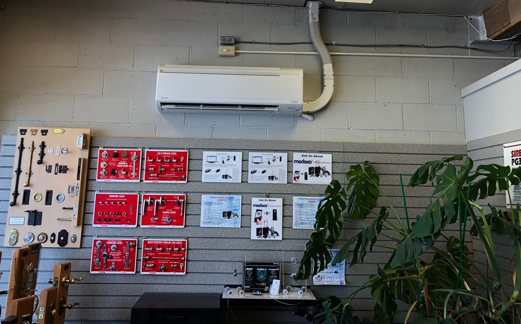 Air conditioner mounted on a gray brick wall with product display boards, a plant, and a small table.