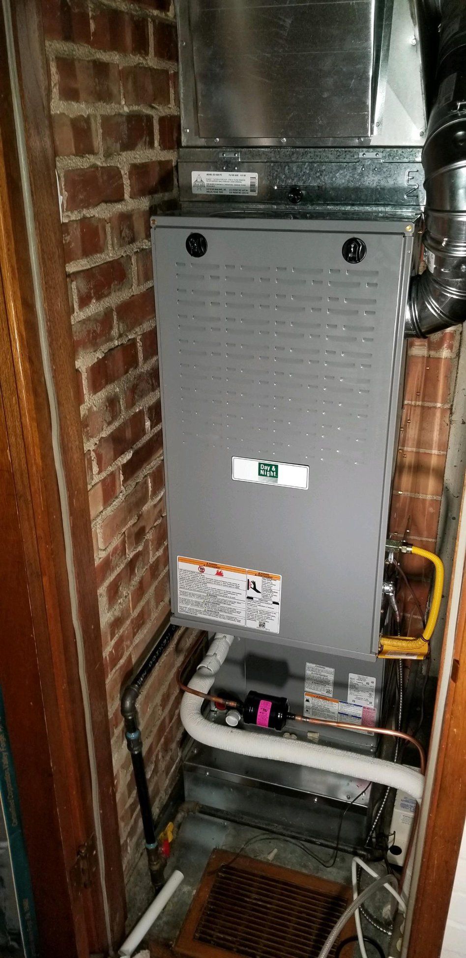 A furnace in a brick-walled enclosure. Gray metal unit with vents, pipes, and warning label.