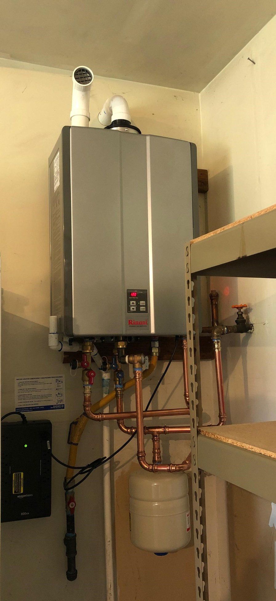 A wall-mounted, silver tankless water heater with copper pipes, a white exhaust pipe, and an expansion tank in a utility room.