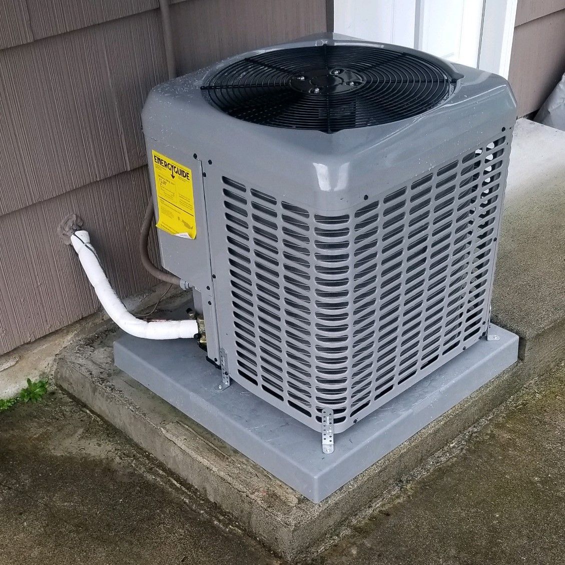 Gray air conditioning unit on a concrete base, mounted next to a house exterior.
