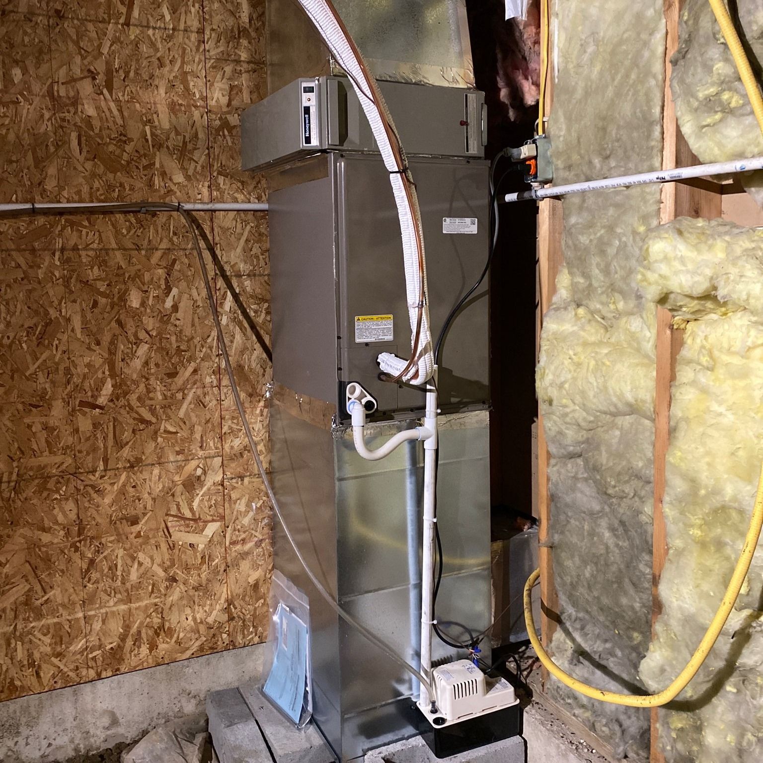 Furnace and ductwork in a utility room. Metal unit is on cinder blocks, next to insulation and OSB wall.