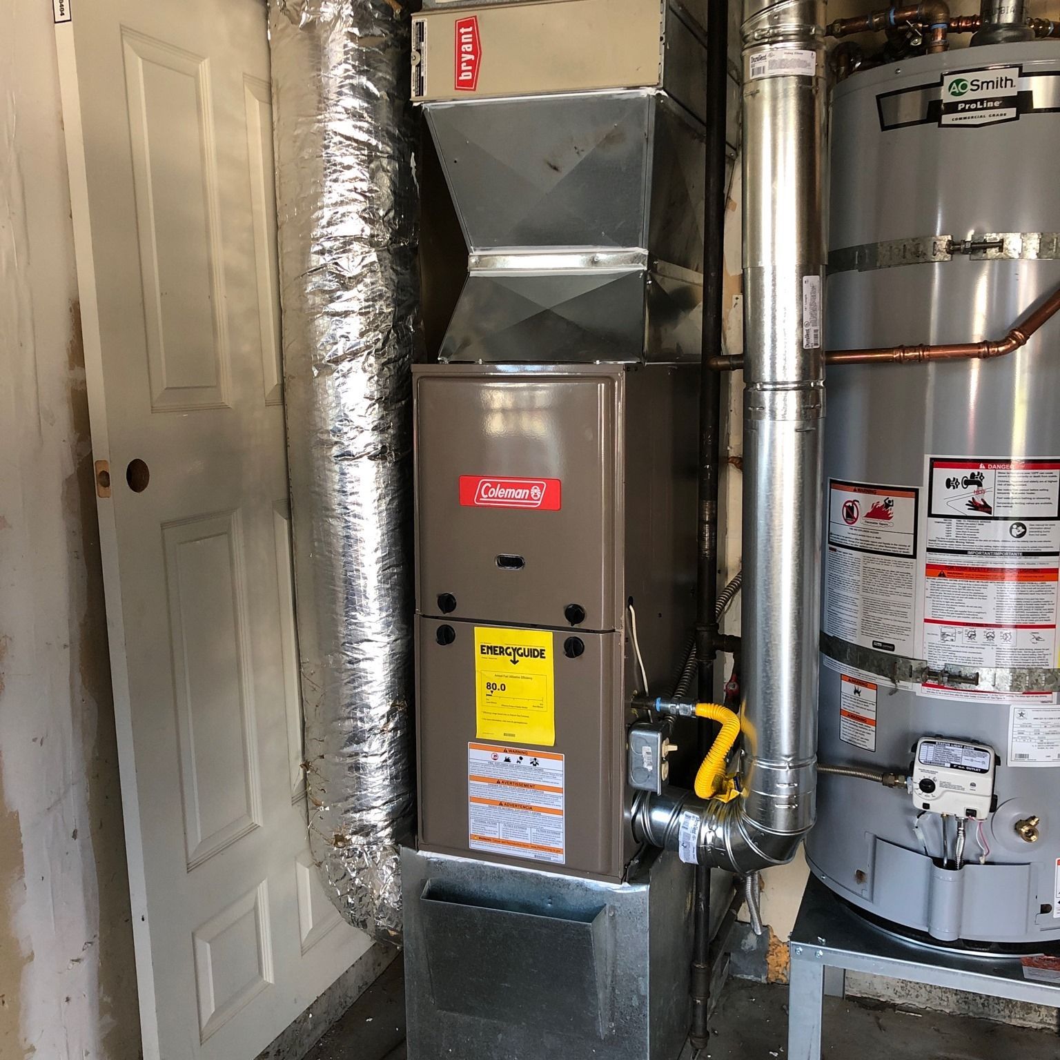 Furnace and water heater in a utility room. Coleman furnace, silver vent pipe, and a white door are visible.