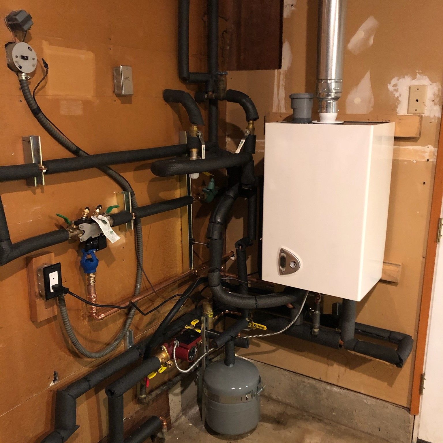 White wall-mounted boiler with connected black pipes and a gray expansion tank in a basement setting.