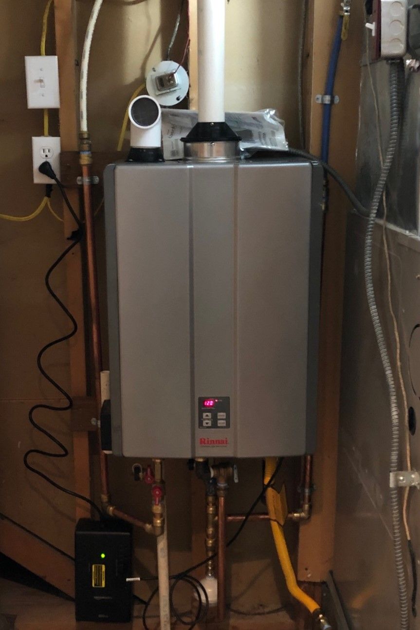 Tankless water heater installed on a wooden wall with copper pipes, a vent, and electrical outlets.