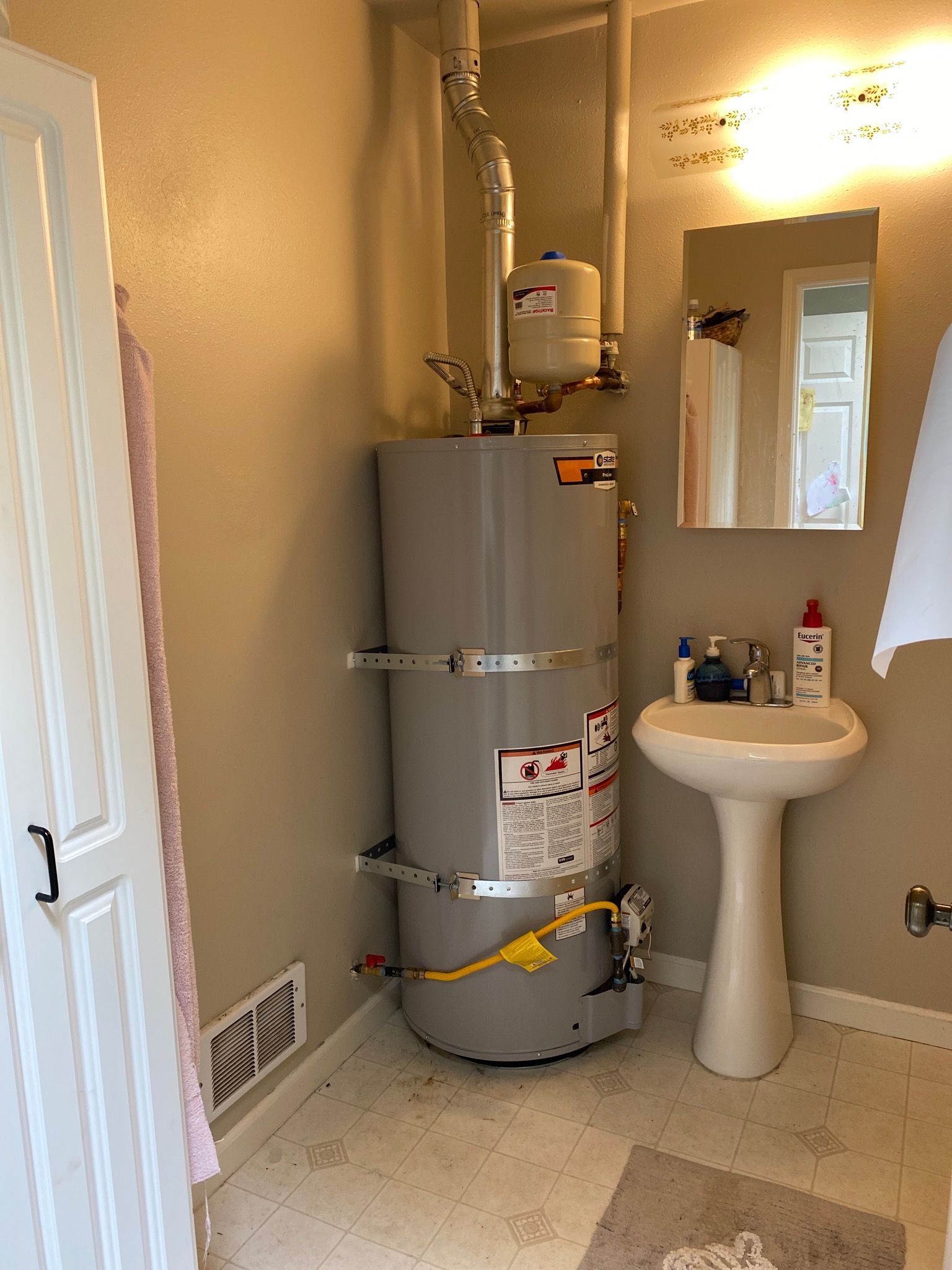 Bathroom with water heater, pedestal sink, mirror, and cabinet. Neutral-colored walls and flooring.
