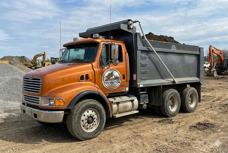 An orange dump truck parked on a construction site with a gray cargo bed loaded with soil.
