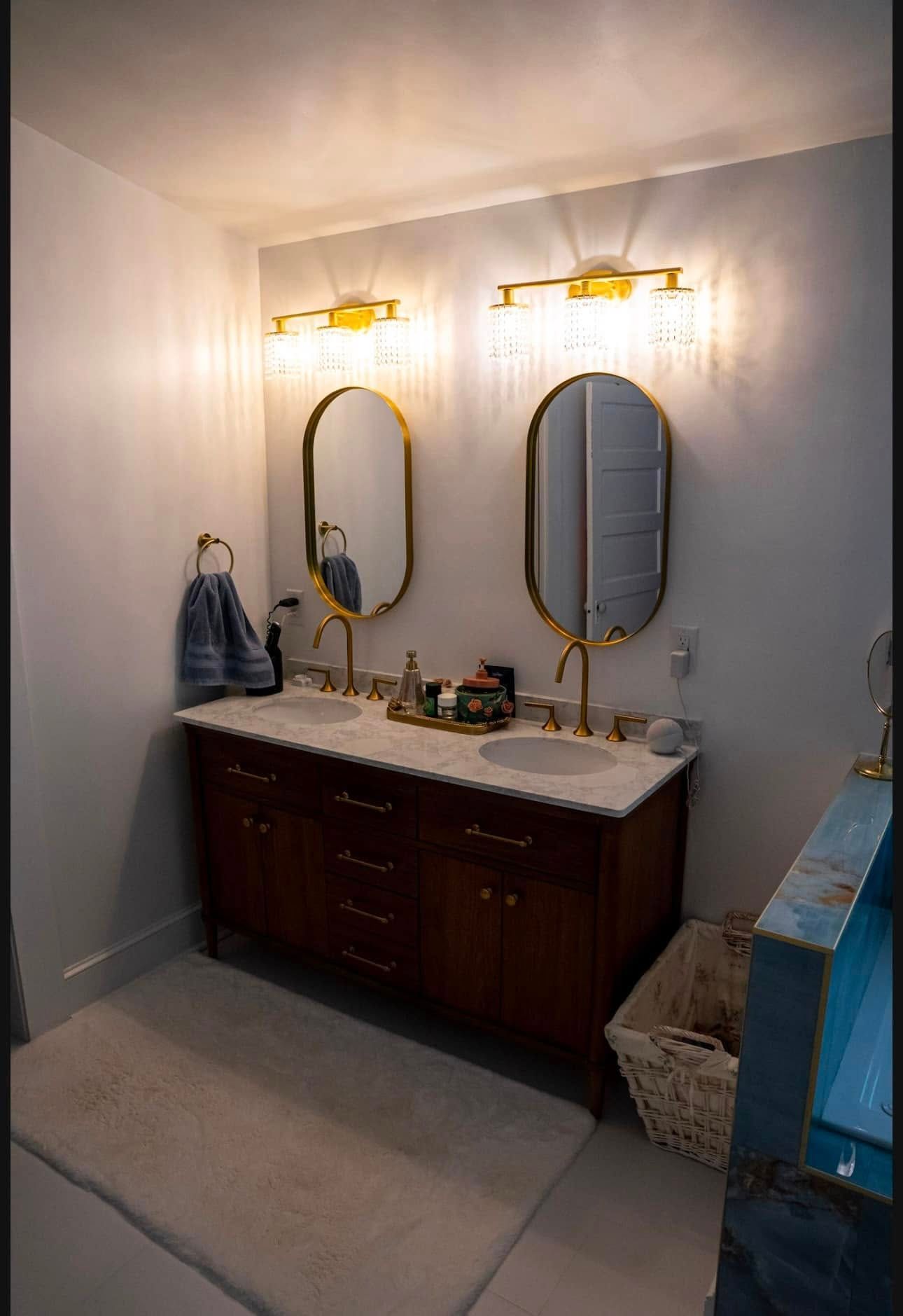 A double vanity bathroom with dark wood cabinets, marble countertop, dual oval gold mirrors, and gold crystal light fixtures.