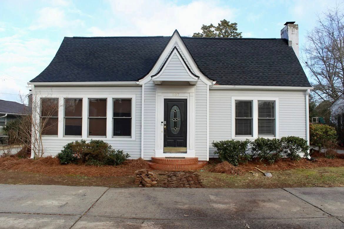 A single-story white house with a black shingled roof, a central gabled entryway, and a driveway in the front.