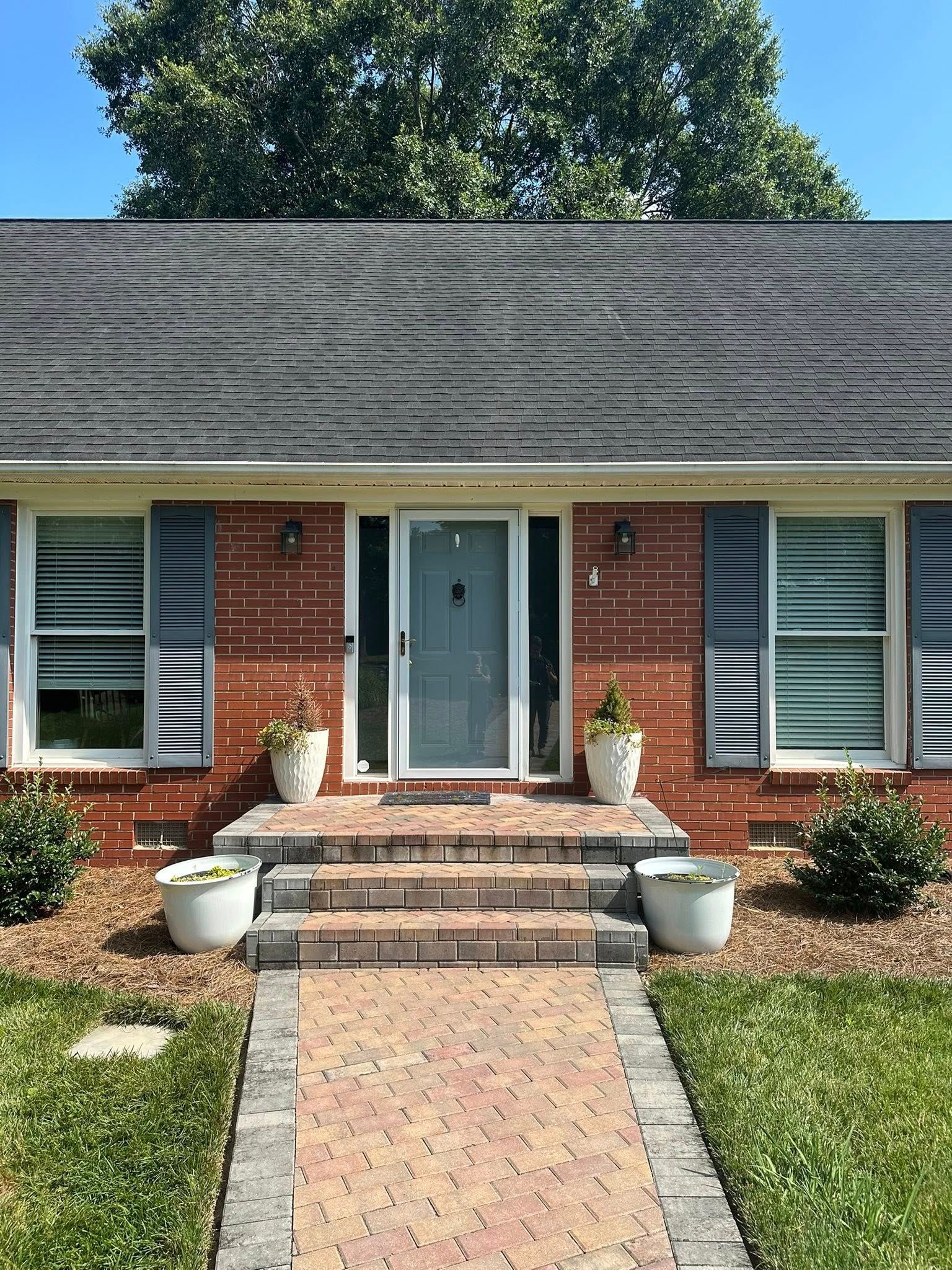 A brick house front with grey shutters, a grey front door, a brick walkway, and two white potted plants by the steps.