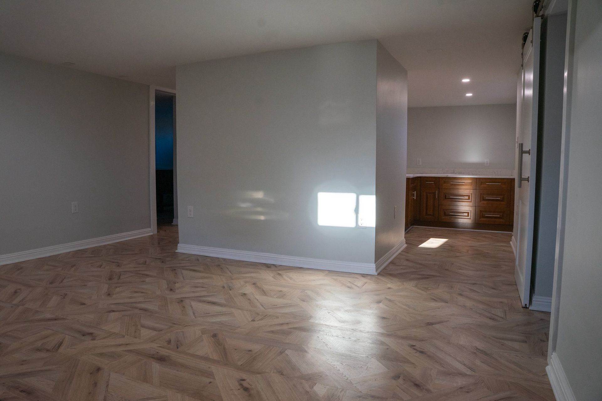 An empty room with light wood-patterned floors, grey walls, and a recessed area containing wooden cabinets.