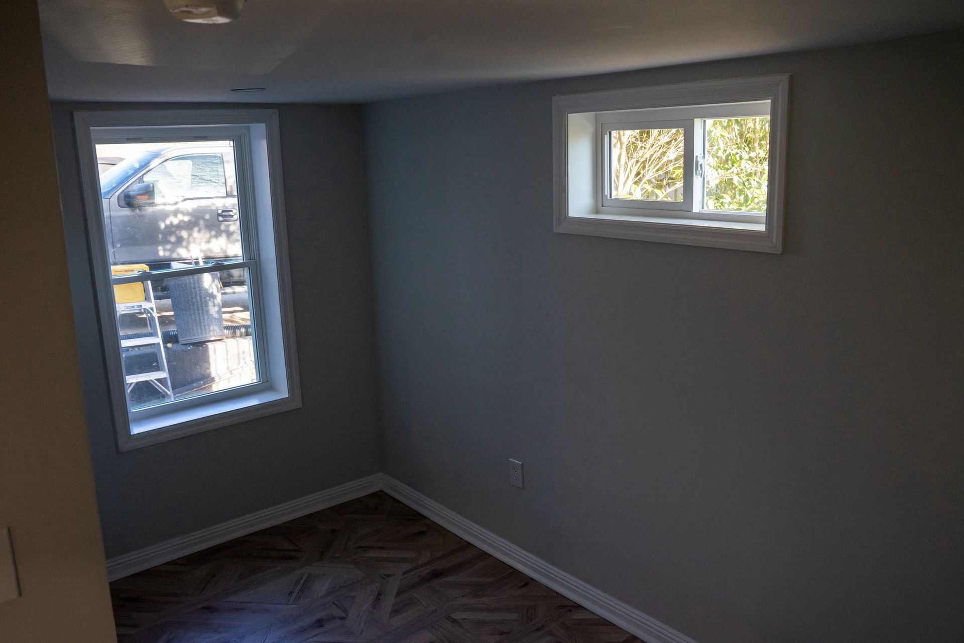 A small room with gray walls, wood-patterned flooring, a tall rectangular window, and a horizontal basement window.