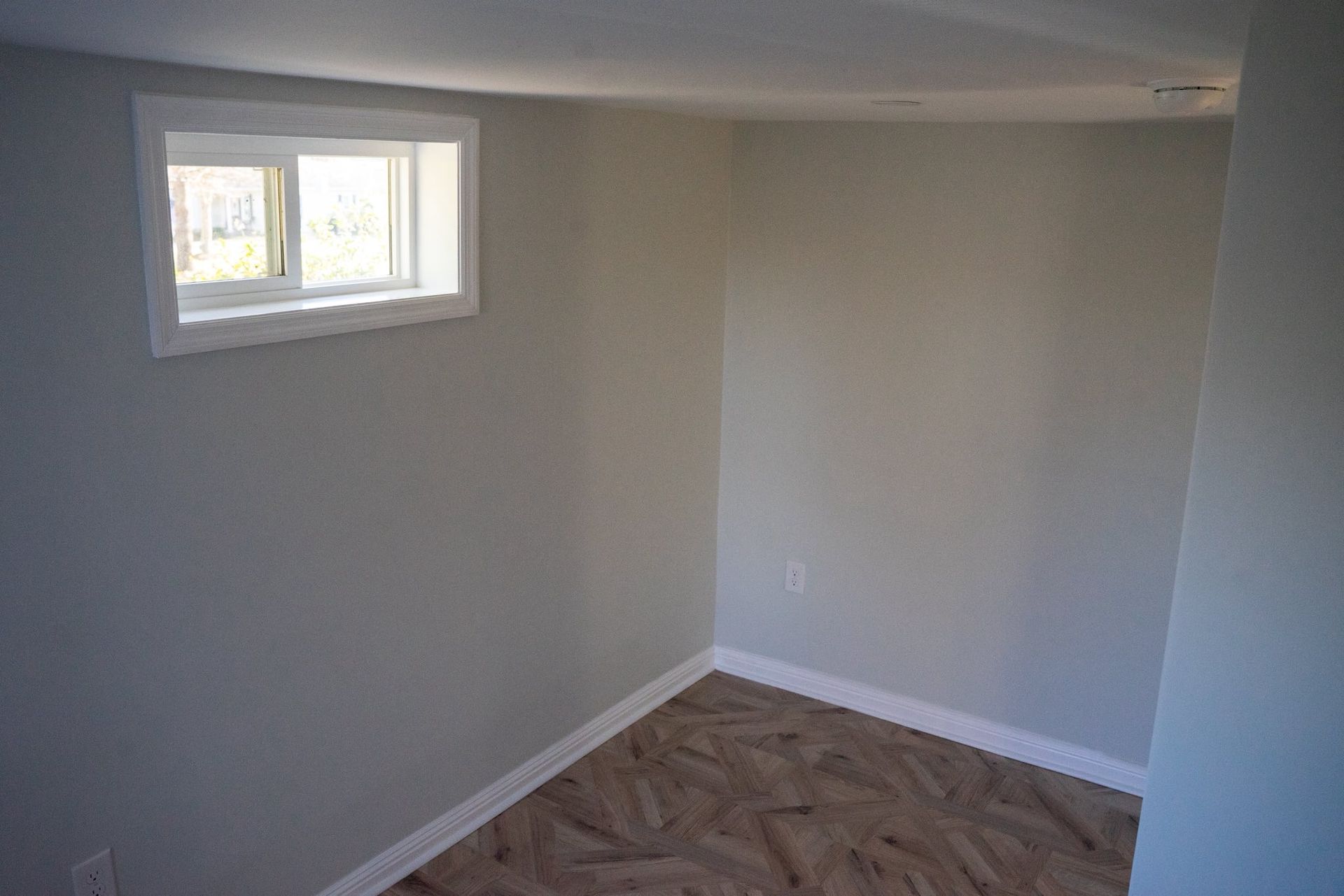 A small, empty room with light gray walls, a white-framed window, and wood-patterned flooring.