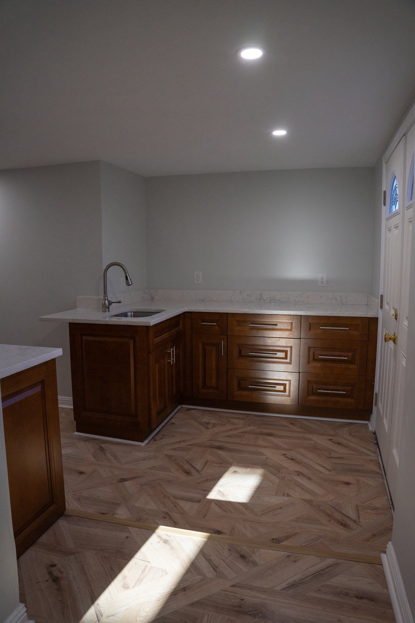A kitchen corner featuring wooden cabinets, a sink, white countertops, and light-colored herringbone flooring.