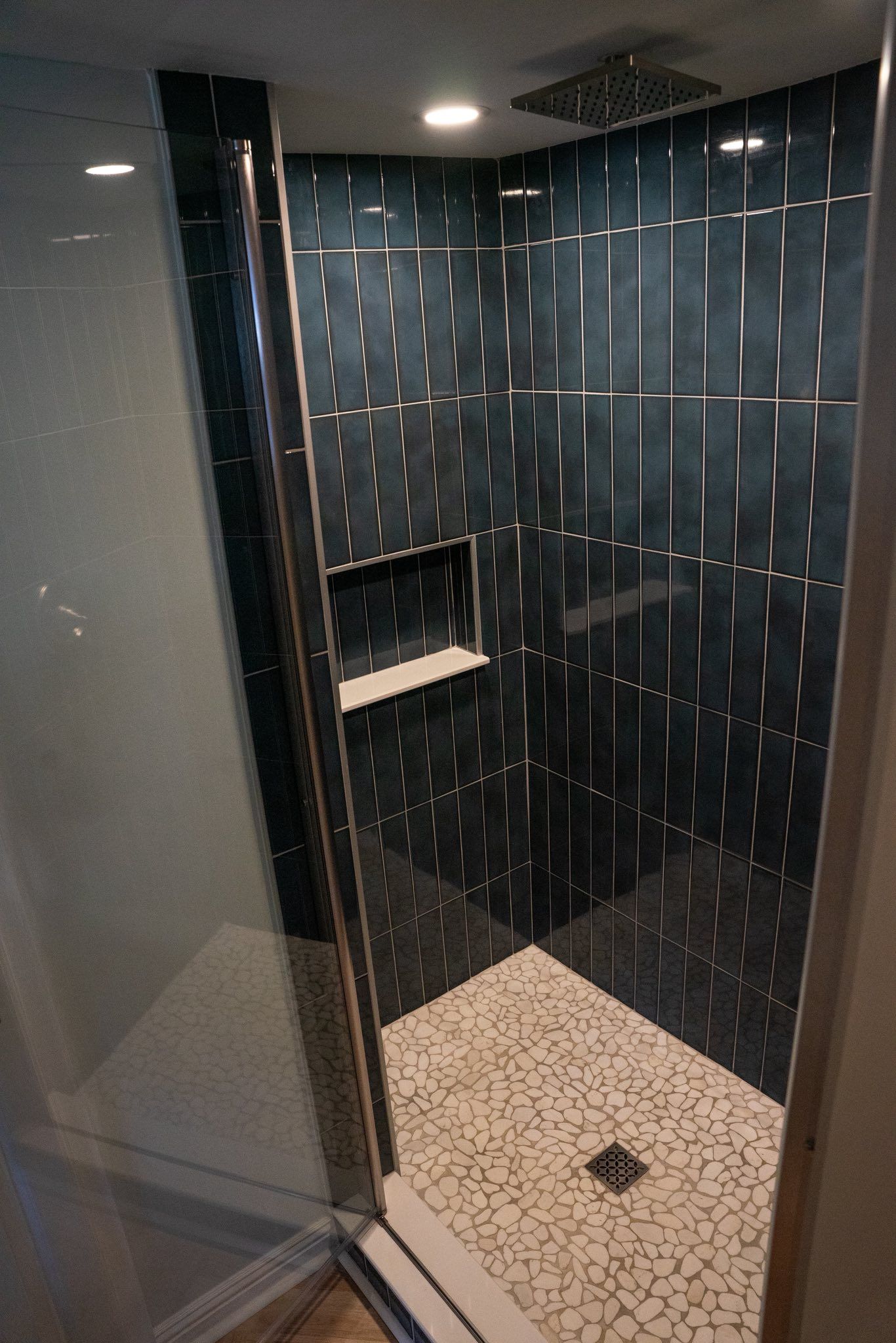 A walk-in shower with dark blue vertical tiles, a built-in recessed shelf, and a light-colored pebble floor.