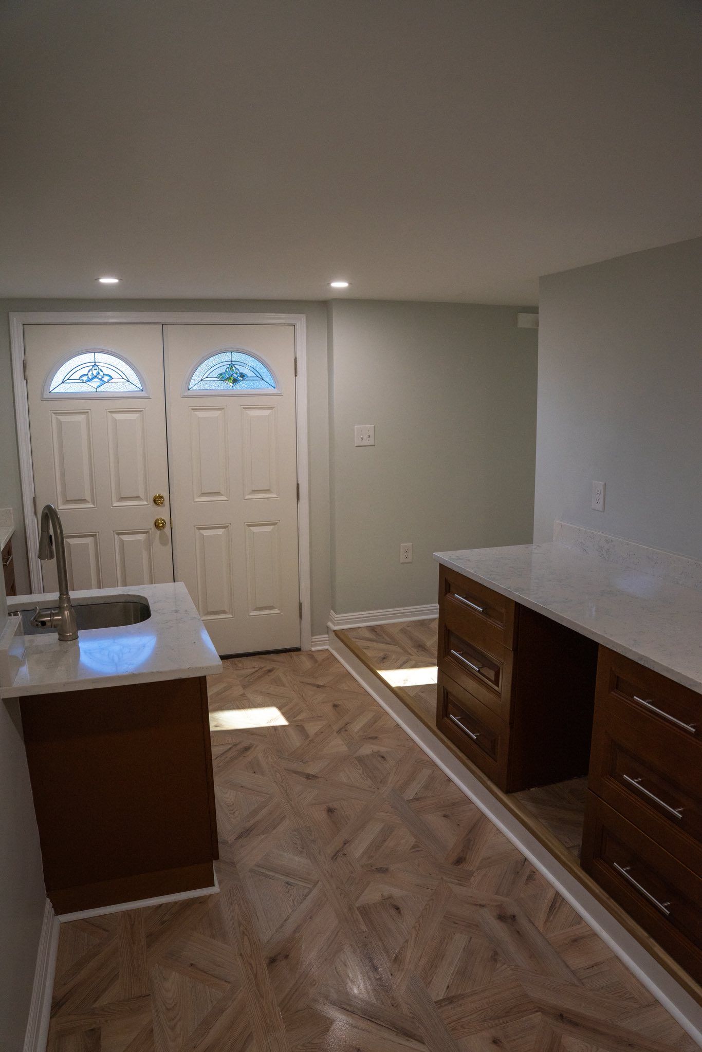 A kitchen area with brown cabinets, white countertops, and wood-patterned flooring leading to white double doors.