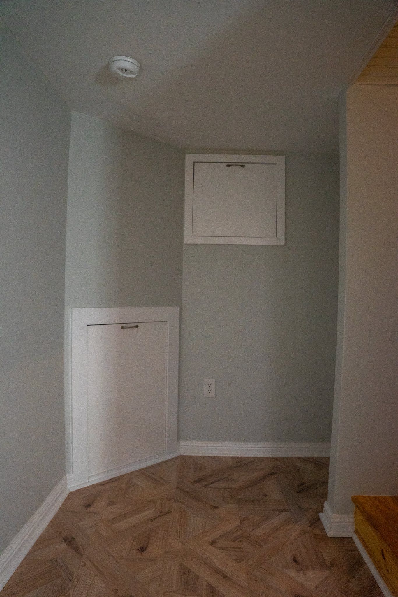 A corner of a room featuring light grey walls, herringbone wood flooring, and two white built-in access panels.