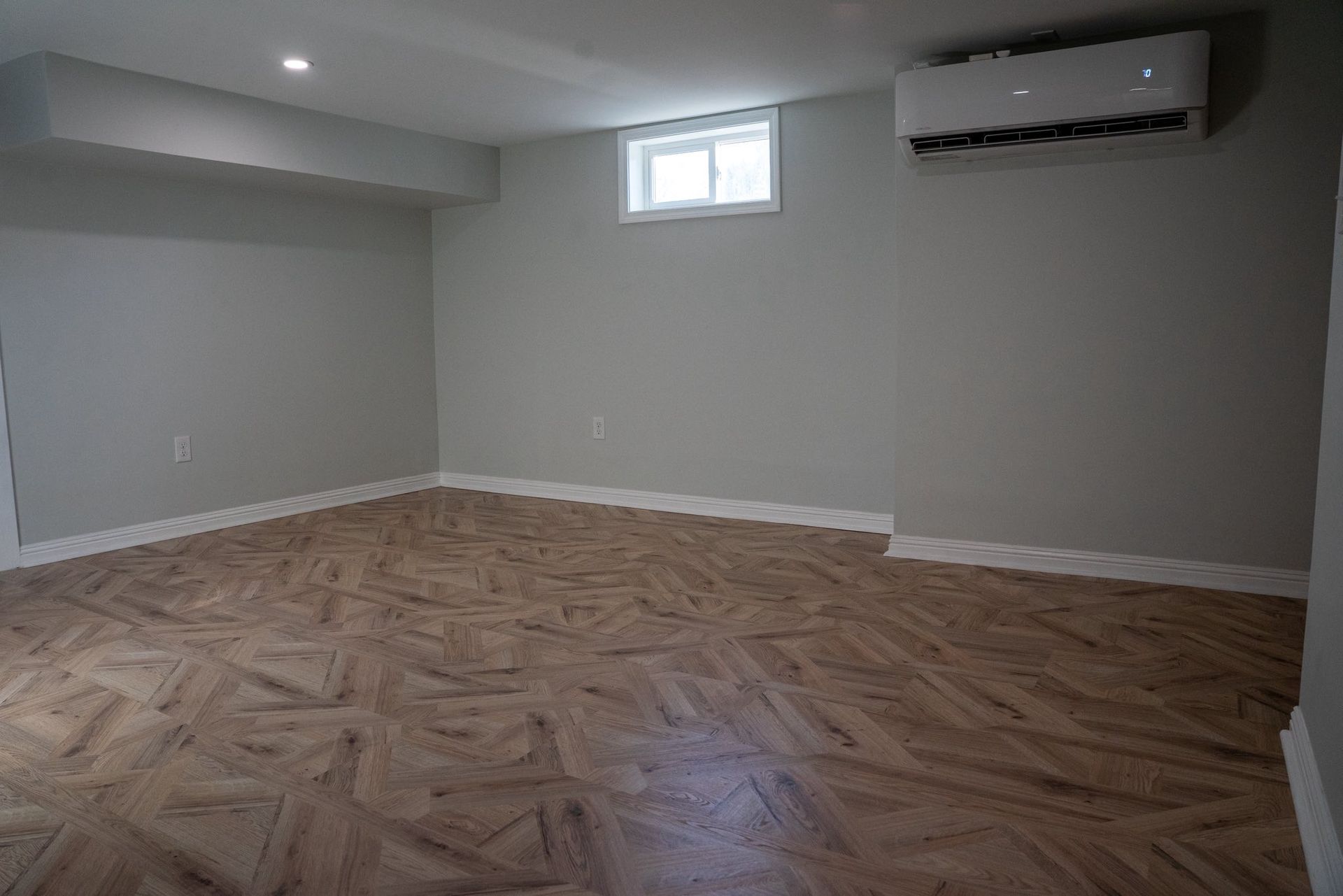 A modern, empty room with light gray walls, wood-patterned flooring, a small window, and a wall-mounted AC unit.